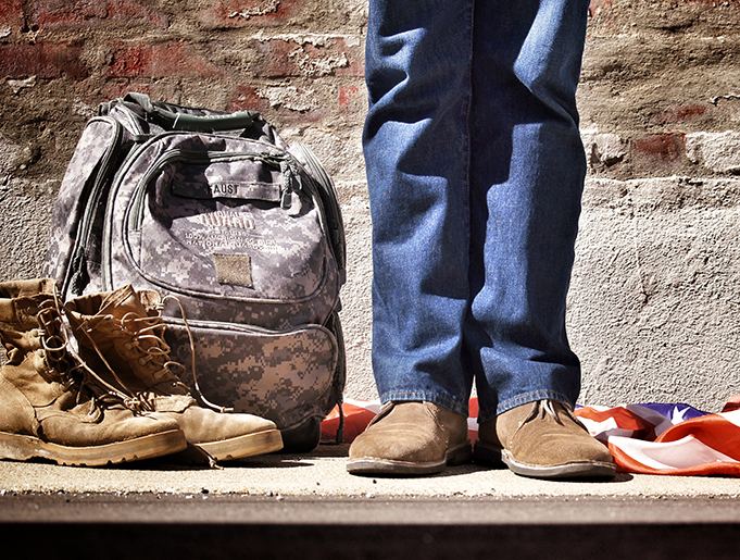 Person wearing jeans and boots standing next to worn backpack and red fabric against brick wall outdoors