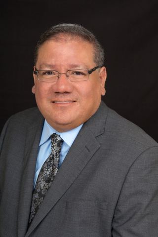 Professional headshot of middle-aged man in gray blazer and blue tie wearing glasses against dark background.