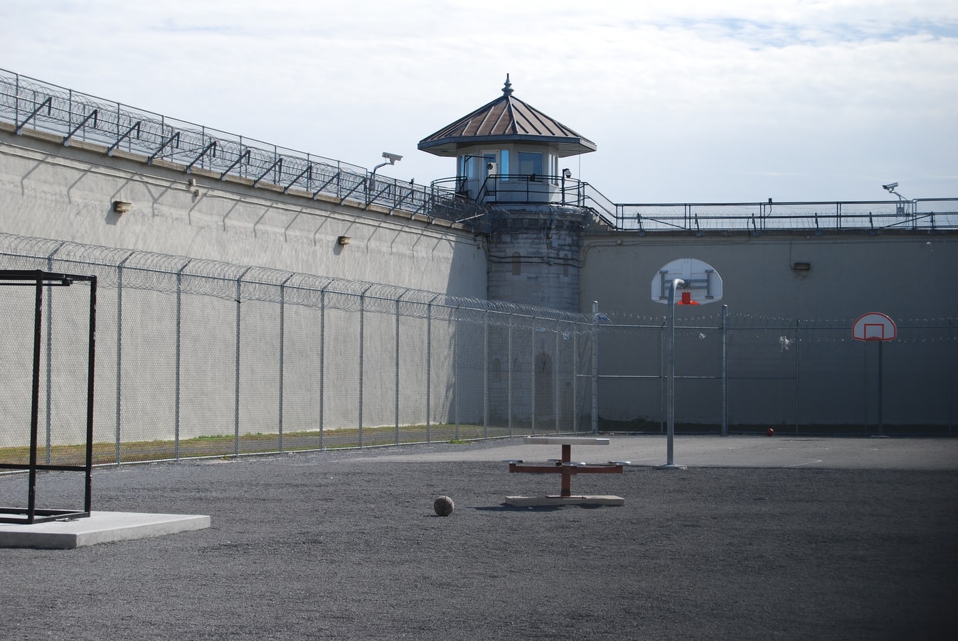 Prison yard with guard tower, basketball hoop, and recreational facilities enclosed by barbed wire fencing.
