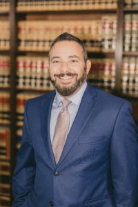 Professional man in blue suit and tie smiling in front of law library bookshelves with legal books