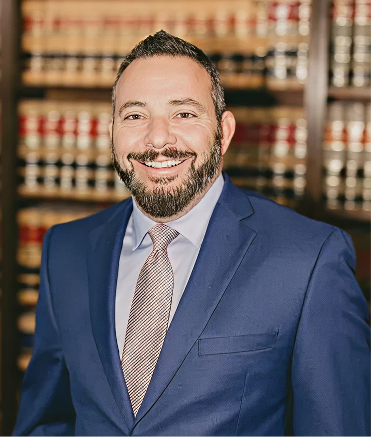 Professional headshot of smiling male lawyer in blue blazer and tie in front of law library bookshelves.