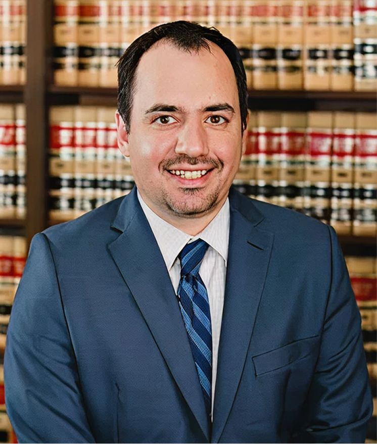 Professional headshot of male attorney in blue suit and striped tie standing in front of law library bookshelves.