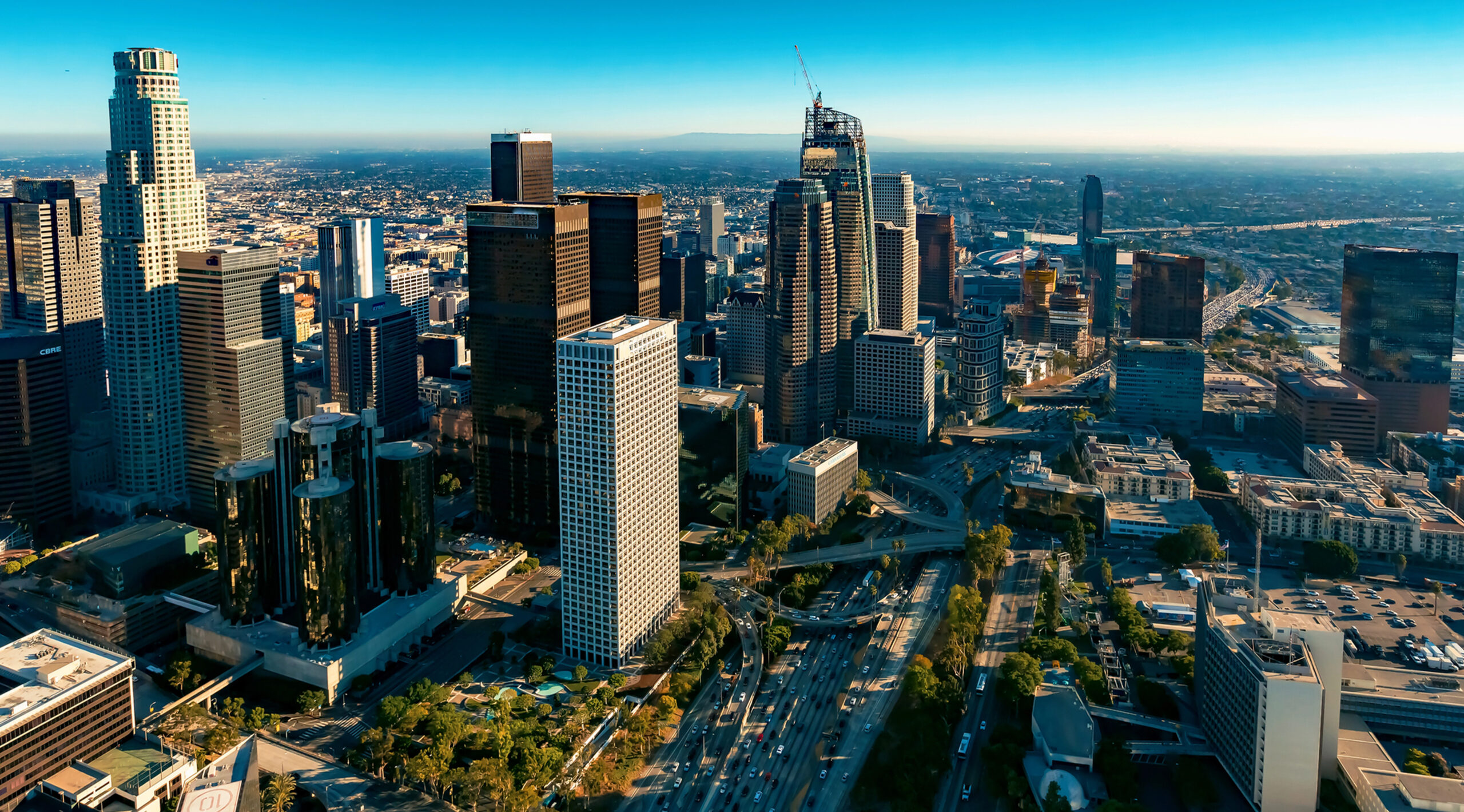 Aerial view of downtown Los Angeles skyline with high-rise buildings, highways, and mountains in the distance under clear
