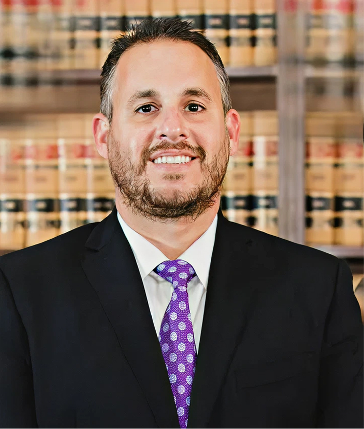 Professional portrait of a male attorney in black suit and purple tie with law books in background.