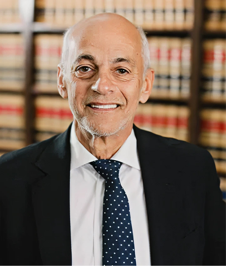 Professional headshot of bald man in suit and tie smiling in front of law library bookshelves