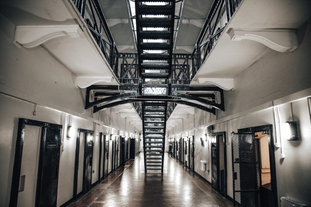 Modern prison cell block with symmetrical rows of doors, metal railings, and LED ceiling lights in a long corridor.
