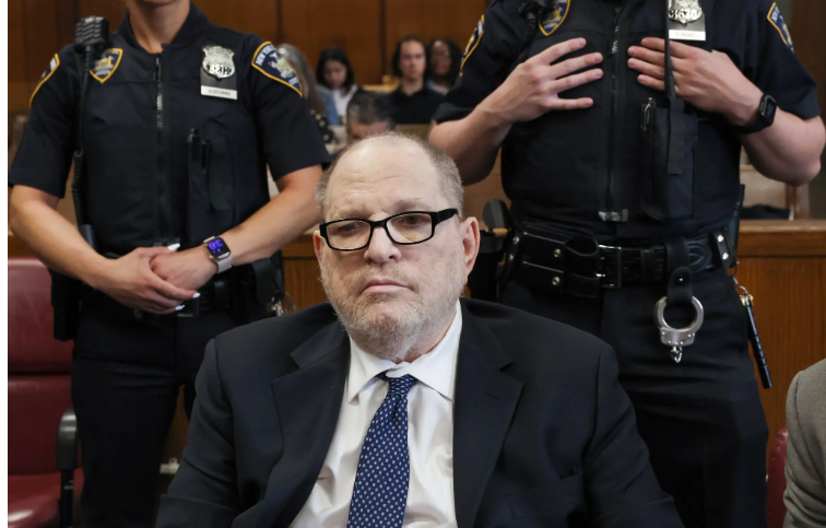 Man in suit and glasses sitting in courtroom with police officers standing behind him